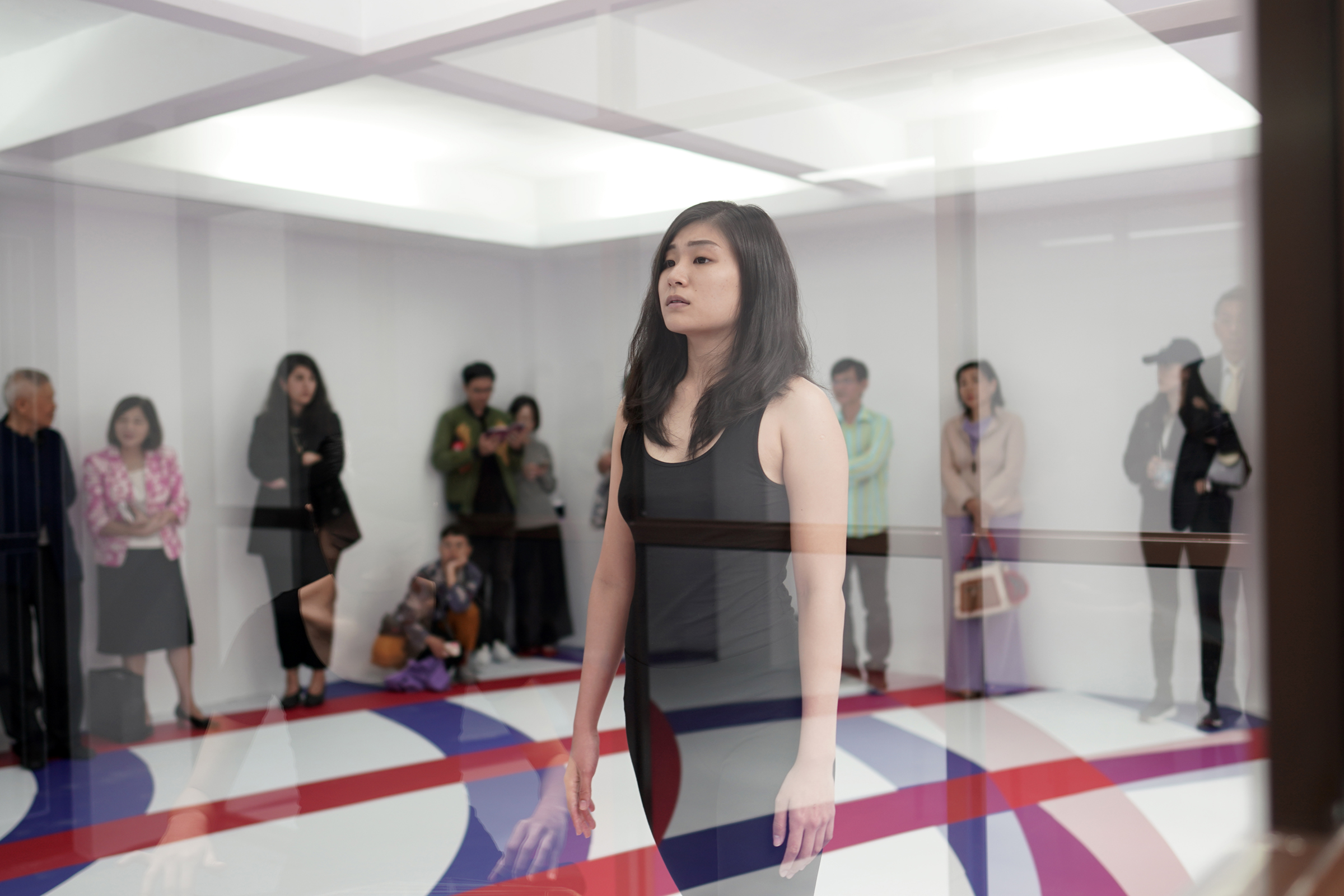 A performer walking towards the left, behind glass and audience in the background