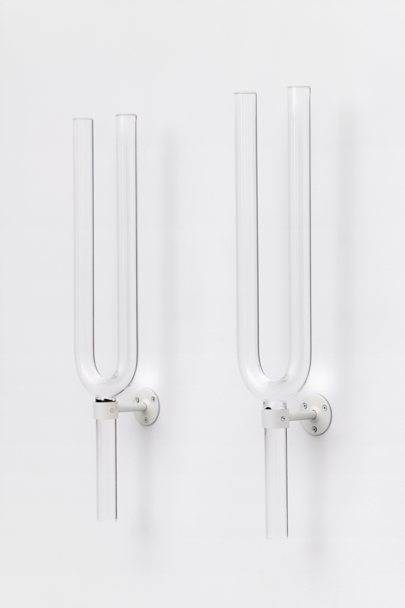Close up of two glass tuning forks on a white wall.